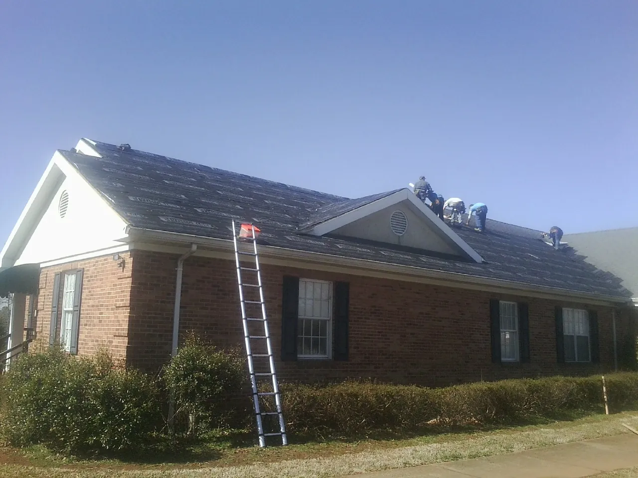 Crew tearing shingles off a brick ranch roof with a ladder set — Emergency Roof Repair work by our crew in De Soto